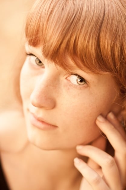 A close-up shot of a woman's face showing her natural skin texture, including freckles and slight imperfections. She is smiling gently at the camera, exuding confidence and self-acceptance. Natural light is illuminating her face, enhancing her natural beauty.