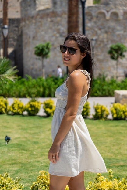 A young woman wearing a short, floral print sundress from Shein, paired with white sneakers and sunglasses, standing in a park on a sunny day.