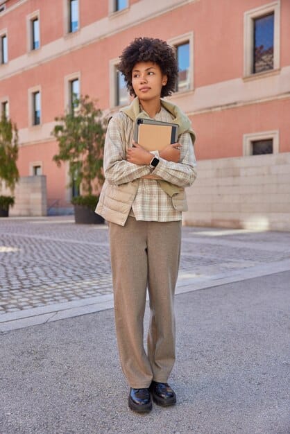 A young person, mid-20s, with dark, curly hair, dressed in a muted plaid blazer, a dark turtleneck, and round framed glasses, holding a worn book. They are seated in an antique library setting, bathed in soft, warm light, embodying the 'Dark Academia' aesthetic.