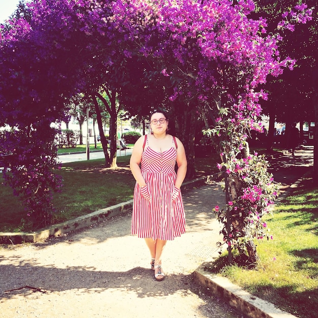 A fashion influencer stands casually in a serene park, wearing a stylish, brightly colored dress. Soft sunlight filters through the leaves, creating dappled shadows. The background is slightly blurred, emphasizing the outfit.