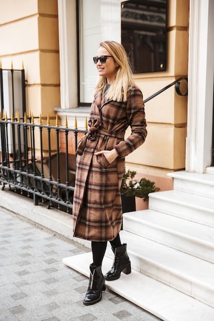 A woman wearing a beige trench coat over a simple black dress and ankle boots, walking down a city street. The outfit epitomizes the effortless elegance of The 2025 Guide to Minimalist Fashion: Less is More.