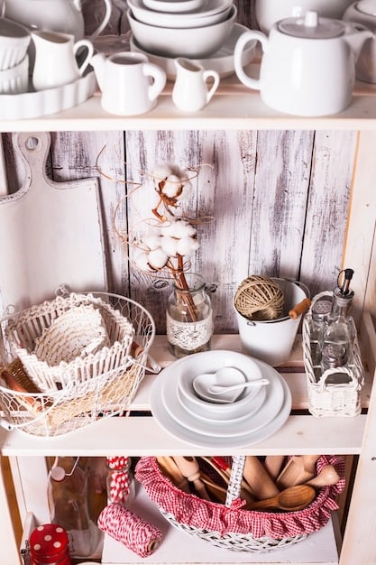 A kitchen designed in the cottagecore style, featuring open shelving with antique dishware, a farmhouse sink, and a gingham patterned curtain. Natural light filters through the window, highlighting the wood textures and the overall rustic warmth that contributes to Will the popularity of cottagecore decor continue its 20% annual growth into 2025?.