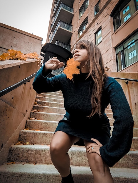 A street style shot featuring a woman wearing a fall-inspired outfit. She is wearing a deep teal coat, a rust orange scarf, and olive green boots. The photo is taken in an urban setting with autumn foliage in the background. The image is designed to showcase how to incorporate the top 5 trending colors for fall 2025, according to Pantone, into a stylish and wearable outfit.
